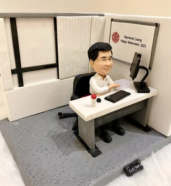 一件為上司Raymond Leung訂製的人像公仔退休禮物。公仔面帶微笑坐在微縮的辦公桌前，桌上有電腦和鍵盤，背景的白板上寫著『Happy Retirement 2021』的祝福語。