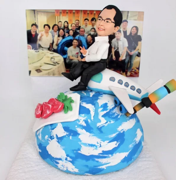 一個充滿心思的榮休禮物人像公仔。模型主角是一位戴眼鏡的男士，他面帶微笑、比著V字手勢，輕鬆地坐在一架環繞地球的飛機上。模型的背景是一張真實的團隊大合照，象徵著美好的工作回憶。地球上還有代表主角興趣的刺身和畫筆模型，設計極具巧思，是一份完美的退休禮物。