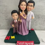 三口之家家庭人像公仔：媽媽穿紫色長洋裝站中間微笑，爸爸著淺灰西裝與領結站右側，孩子穿棕色小西裝站左側比出手勢；三人並肩站在深綠色草地質感底座上，周圍點綴紅色與黃色小花。前方紅色名牌寫有「CuteFigureHK」，背景為水泥紋灰牆與白色檯面。整體高雅溫馨，適合作為家庭人像公仔紀念禮物。