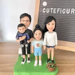 家庭人像公仔四人組：爸爸抱著穿深藍運動套裝的小朋友男孩站在左側；媽媽穿白色上衣與牛仔短褲、綠色鞋在右側；中間是紮高丸子頭、穿淺藍上衣與短褲的小女孩。四人站在草綠色不規則底座上，右前方放著棕色小籃子。背景為木質桌面與立式字母佈告板，上有「CUTeFIGUR…」字樣。整體清新溫馨，展現一家人的日常氛圍，適合作為小朋友主題的家庭人像公仔禮物與居家擺設。