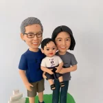 三口之家家庭人像公仔：灰髮戴眼鏡的父親穿藍色T恤與卡其短褲，母親留及肩黑髮穿灰色T恤與深色長褲，兩人相擁站立，中間懷抱一名穿白色上衣、深色短褲與黑色小鞋的嬰兒公仔；三人微笑表情自然，立於綠色草地樣式的底座，背景為簡潔淺色牆面。整體清新溫馨，適合作為新生兒百日宴的家庭人像公仔禮物。