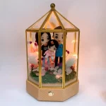 八角金色玻璃展示燈盒中的家庭人像公仔場景：爸爸、媽媽與小女孩站在綠色草地與粉色花瓣點綴的地台上，周圍有白色花朵與小型植栽裝飾；盒內串燈發出暖黃色光，映照三人溫馨表情與服飾（媽媽黑色上衣、爸爸藍色外套、小女孩粉色系穿搭）。底座為木紋質感並設有按鈕。整體夢幻浪漫，適合作為百日宴家庭人像公仔禮物與夜燈擺設。