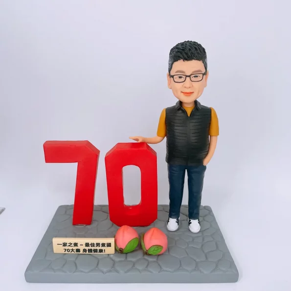 一個戴眼鏡、身穿黃衣黑色背心的人像公仔，站在灰色地台上，旁邊有大型紅色「70」字樣和兩個壽桃，前方有祝福字牌。