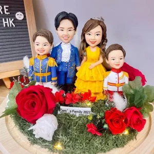 四口之家主題家庭人像公仔擺設：爸爸穿藍色王子禮服、媽媽穿亮黃色層次長裙並配髮飾，兩名孩子分別穿藍金王子服與白紅小王子服。公仔站在苔蘚草地造景上，周圍點綴紅白玫瑰與小燈串，前方有「Family 2022」銘牌。背景為木紋底座與字母板，整體夢幻童話風格，適合作為家庭人像公仔禮物與居家展示。