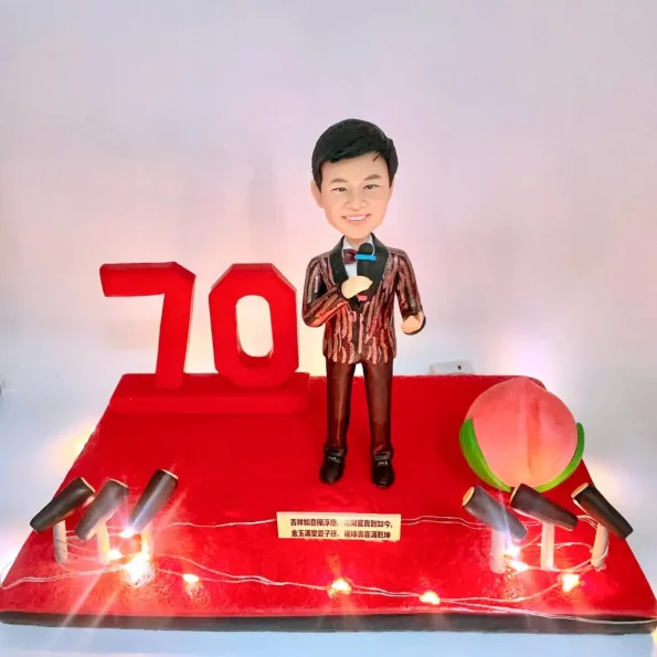 一個穿著西裝、手持麥克風的人像公仔站在舞台上，旁邊有紅色「70」字樣和壽桃，舞台上有多支麥克風，底座有祝福字牌。