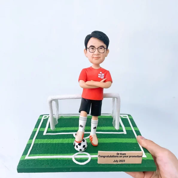 為慶祝Dr Kam升職而訂製的人像公仔禮物。公仔身穿紅色足球服，自信地踩著足球，站在一個微縮足球場的龍門前，前方的名牌寫著「Congratulations on your promotion!」。