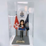 一個裝在透明展示盒中的警察晉升賀禮。禮物主體是一個女性警官的人像公仔，她正站在記者會的講台前，身後是香港區旗、警隊旗幟及「屯門重案第二隊」的背景板，前方的銘牌標明這是祝賀其榮陞警司的禮物。