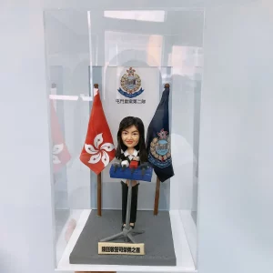 一個裝在透明展示盒中的警察晉升賀禮。禮物主體是一個女性警官的人像公仔，她正站在記者會的講台前，身後是香港區旗、警隊旗幟及「屯門重案第二隊」的背景板，前方的銘牌標明這是祝賀其榮陞警司的禮物。