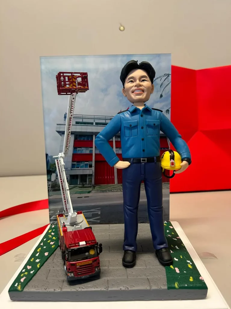一個為一位消防人員訂製的場景模型。模型主角是一位面帶開朗笑容的男性公仔，他身穿藍色工作制服，一手叉腰，另一手拿著黃色頭盔。他身旁是一輛升降台消防車模型，其雲梯高高升起，背景是一張消防局的相片。