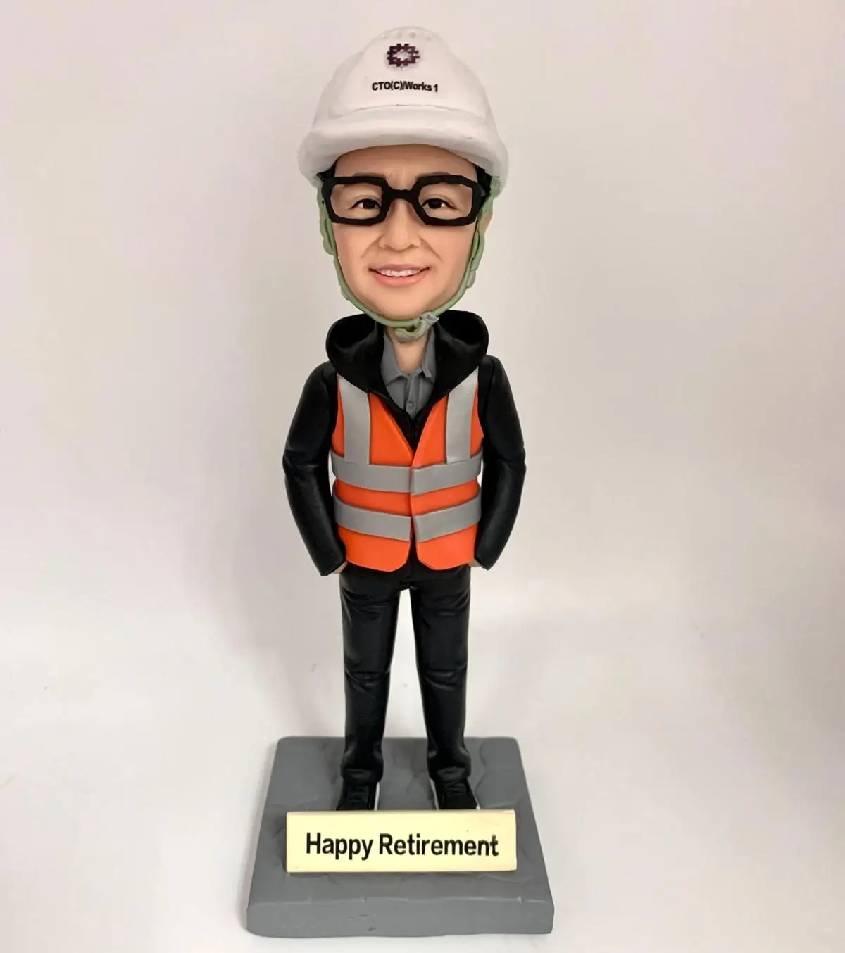 為一名戴眼鏡的工程師所客製的人像公仔，公仔頭戴印有「CTO(C)/Works 1」標誌的安全帽，身穿黑色連帽外套與橙色反光背心，底座上刻有「Happy Retirement」字樣，是一份完美的榮休紀念品。