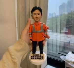 為資深工程師王志華先生訂製的榮休人像公仔，公仔身穿橙色反光外套，其底座刻有「默默耕耘廿四載」的讚頌題字，是一份表彰長期服務的獨特紀念品。
