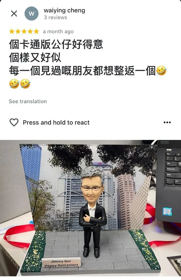 一件為男士「Jimmy Gor」訂製的榮休禮物。一個銀髮、戴眼鏡的男性人像公仔,身穿西裝、雙手抱胸,自信地站立著。背景是一張香港中環金融區的實景照片,以匯豐總行大廈為中心,象徵其事業成就。底座的牌匾寫著「Jimmy Gor Happy Retirement」。