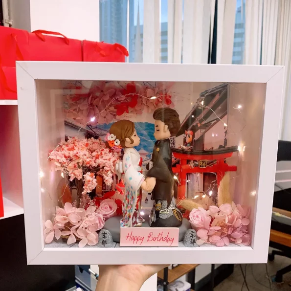 白色相框內的日系情侶場景：女生穿白底彩點和服配花髮飾，男生穿深色和服，兩人面對面牽手站立。背景有紅色鳥居、日式街景與櫻花樹，前景點綴粉色花材與小石碑，上方繞有暖光燈串，底牌寫著「Happy Birthday」。