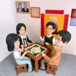 四位家庭成員的人像公仔圍坐在麻雀枱旁，背景有相框、時鐘和揮春，展現家庭一同打麻雀的溫馨時刻。