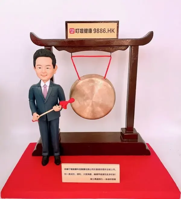 一隻男士人像公仔身穿西裝，手持紅色鼓槌，站在大鑼旁邊。背景設有公司上市敲鑼裝置，上方牌匾寫有公司名稱及股票代碼「9886.HK」。公仔和裝置放置於紅色底座上。這是特別為公司上市設計的人像公仔禮物。