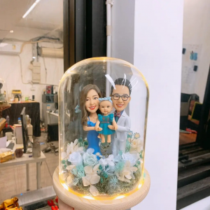 玻璃圓罩中的三口之家家庭人像公仔：媽媽穿藍色洋裝、爸爸穿淺色西裝與眼鏡，抱著戴花環與藍色服飾的嬰孩；底座以苔蘚、牛奶色與藍色永生花環繞，圓罩邊緣有暖黃燈串點綴。整體呈現溫馨典雅與儀式感，適合作為家庭人像公仔禮物與居家擺設。