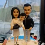 三口之家家庭人像公仔近拍：媽媽留及肩黑髮，穿白色上衣與淺色裙，抱著懷中嬰孩；爸爸穿黑色外套與淺灰長褲站在右側，三人腳穿白鞋並肩而立。公仔置於米白圓形底座，背景為窗外高樓景色與辦公室環境，整體溫馨甜蜜，適合作為家庭人像公仔禮物。