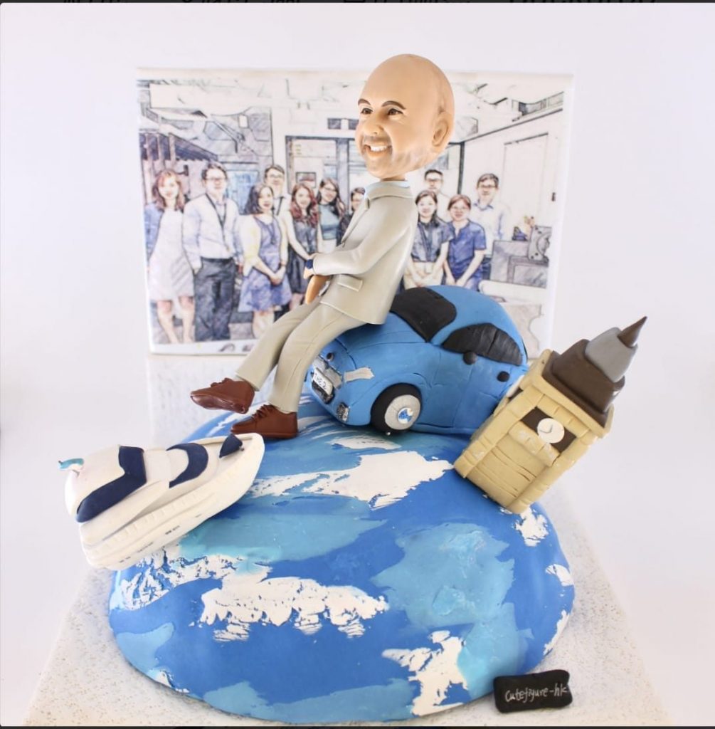 一個精緻的客製化榮休禮物場景模型。模型主角是一位笑容燦爛的光頭男士人像公仔，他輕鬆地坐在一輛藍色汽車上，汽車則停在一個地球模型頂端。地球上還有倫敦大笨鐘和跑鞋模型，象徵著旅行和個人興趣。背景是一幅素描風格的團隊合照，整個作品充滿故事性，是極佳的退休禮物選擇。