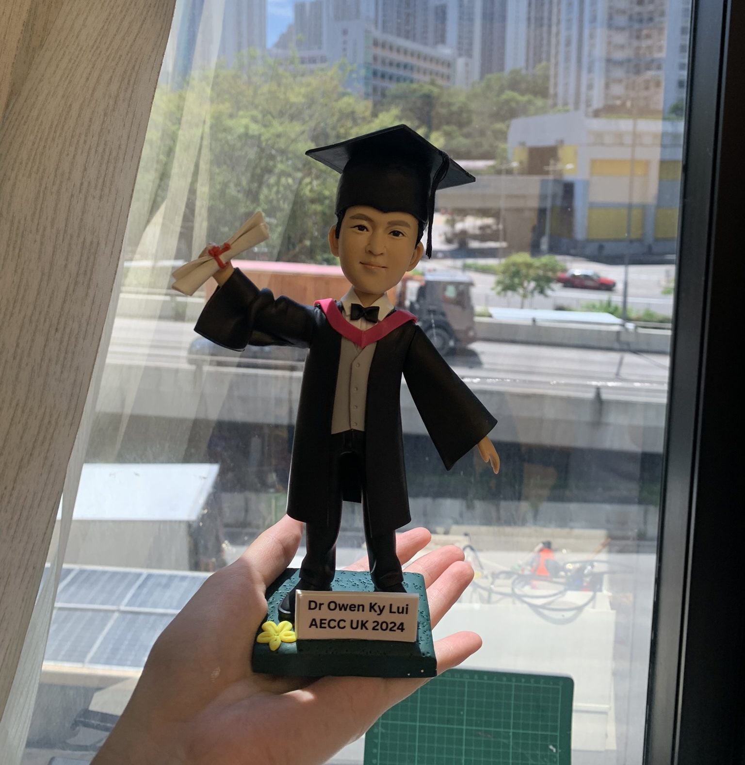 一個男生畢業生人像公仔，穿著學士袍和帽子，手持畢業證書，背景為窗外城市景色。