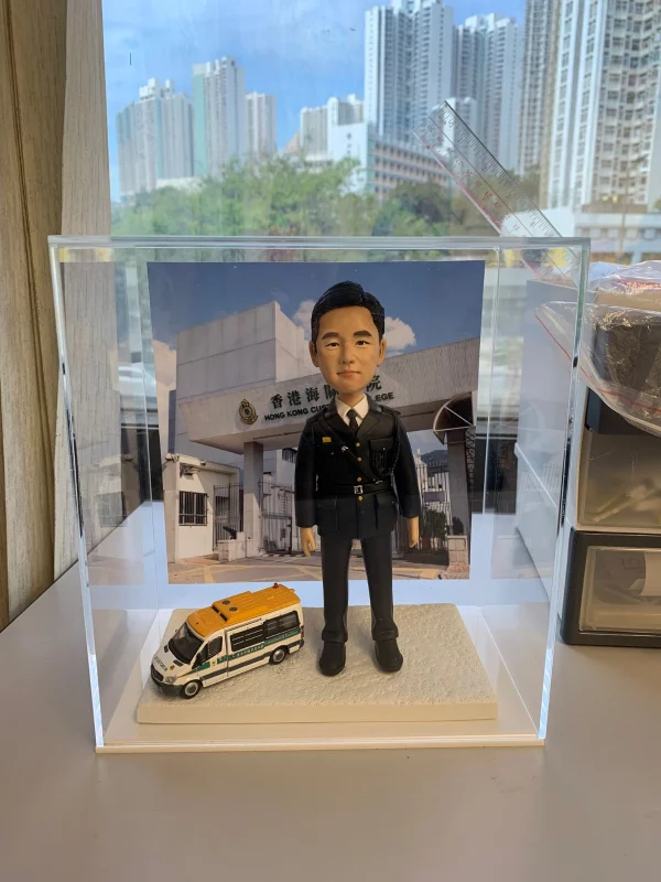 身穿深藍色海關制服的訂造人像公仔，旁邊有海關車模型，背景是香港海關學院，是非常適合送給海關人員的退休禮物或升職禮物。