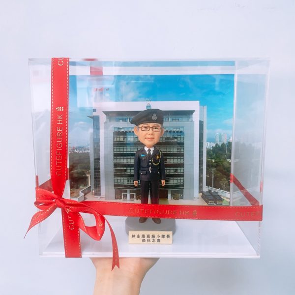 一個身穿深色制服並戴黑色貝雷帽的訂造Q版真人公仔，站在印有建築物背景的透明禮物盒內，是公務員退休禮物或升職禮物的窩心之選。