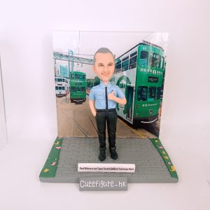 一位身穿淺藍色制服的男士Q版真人公仔，豎起拇指，背景是香港電車軌道和綠色電車，前方有刻字「Best Wishes to our Super Smart & Brilliant Tramways Hero!」，是表彰電車英雄貢獻的紀念公仔。
