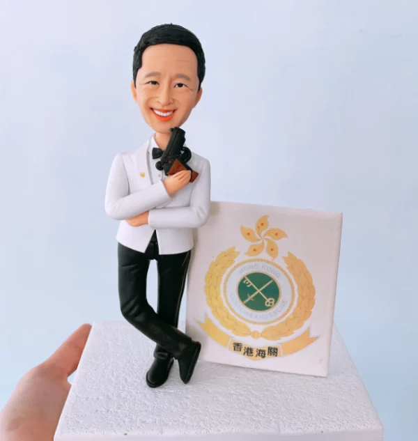 穿白色禮服的Q版真人公仔，手持手槍，靠在香港海關徽章旁，是送給海關人員的特別退休禮物或升職禮物。