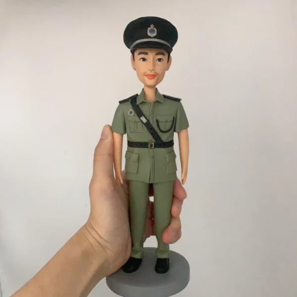 一個穿著綠色公務員制服的訂造人像公仔，戴著黑色帽子及斜孭帶，被一隻手握著底部，是送給公務員的退休禮物或升職禮物首選。