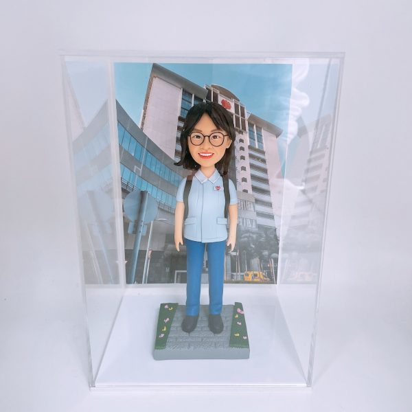 一位背著背包的女性人像公仔，她身穿淺藍色制服，站在鋪有落花的石板路上，背景為仰視角度的現代化醫院或機構大樓照片，是一款象徵開啟新旅程的榮休禮物。