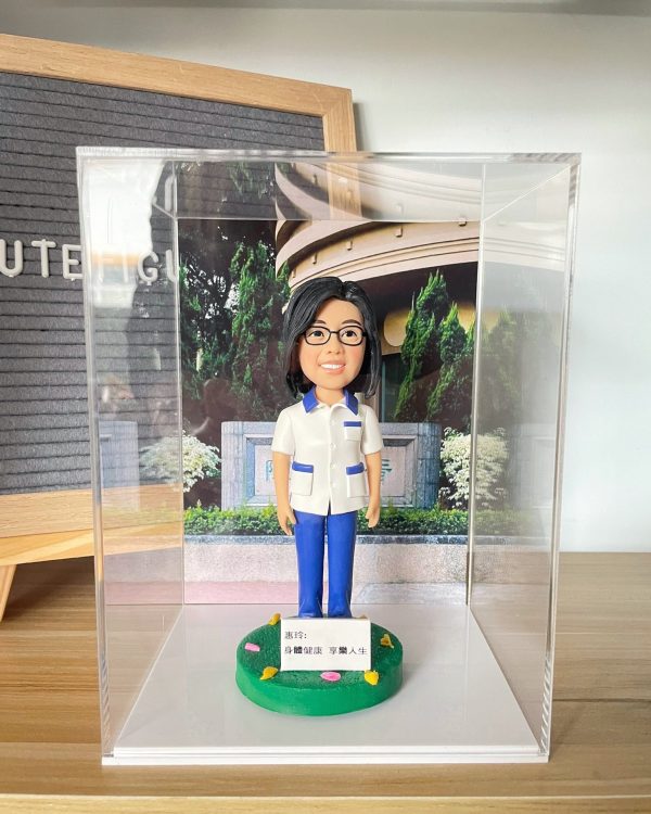 一位身穿白色及藍色制服的女性醫護人員人像公仔，被放置在透明展覽箱中，背景為其工作機構的入口照片，綠色草地底座上立有「身體健康 享樂人生」的祝福牌，是一份意義深遠的榮休禮物。