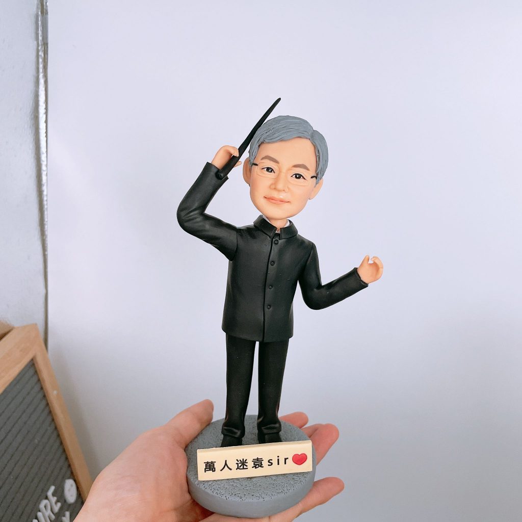 一位銀髮男老師的人像公仔,他身穿黑色中山裝,手持指揮棒,姿態如同樂團指揮。底座的銘牌上寫著「萬人迷sir」,是一款充滿愛戴之情的榮休禮物或紀念品。