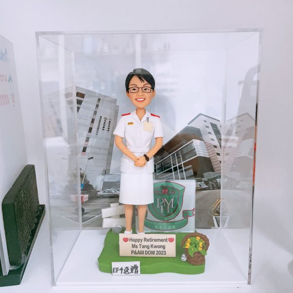 為瑪嘉烈醫院（PMH）P&AM 部門主管 Ms Tang Kwong 製作的榮休人像公仔，公仔身穿護士長制服，背景為醫院大樓，底座上有高爾夫球桿、金色果實籃及寫有退休祝福的牌匾，並註明由「EF仕送贈」。