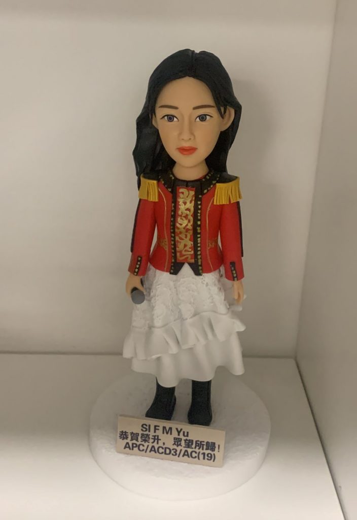 一隻女性人像公仔，穿著紅色金邊肩章外套和白色多層次裙，手持麥克風，站在白色圓形底座上。底座前有一塊寫有祝賀字句的牌。這款人像公仔以特別造型設計，適合做升職禮物。