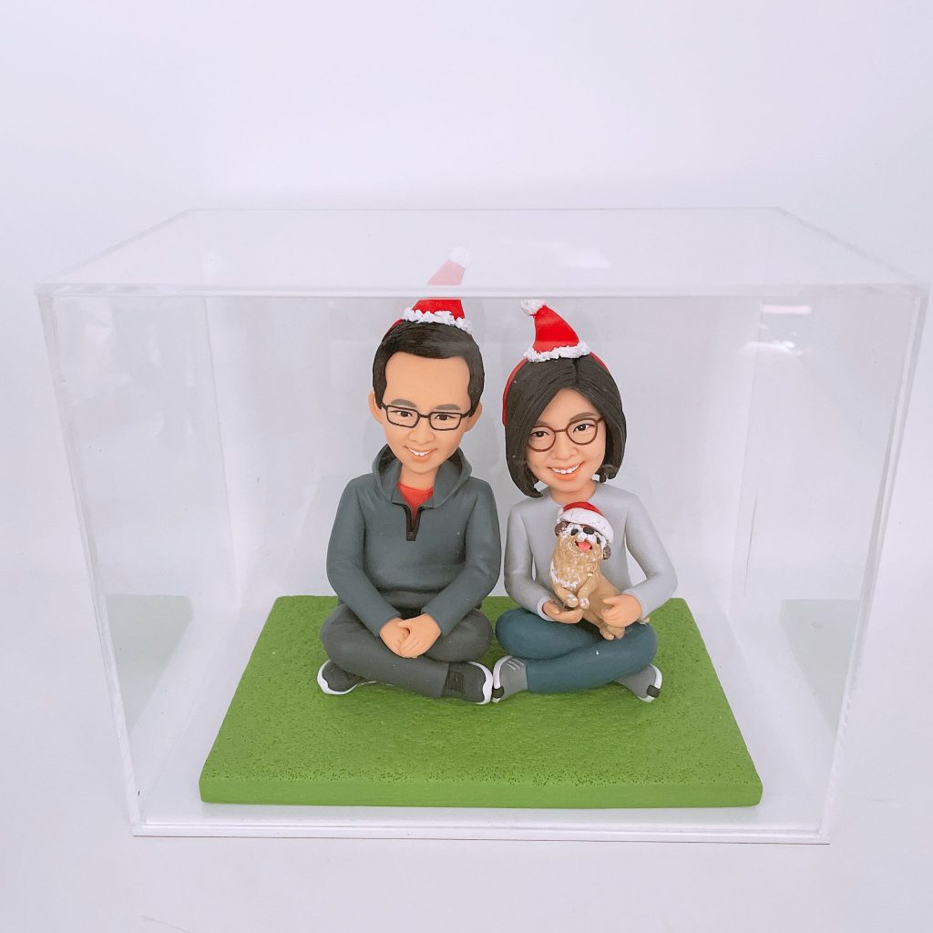一對情侶和一隻狗的人像公仔，頭戴聖誕帽，坐在綠色草地造型底座上，裝在透明盒內。