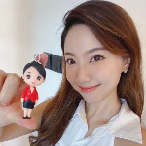 一位氣質優雅的女士，手持一個根據她樣貌製作的人像公仔鑰匙扣，公仔身穿紅黑色職業套裝，笑容甜美，是一款精緻便攜的職場紀念品或榮休禮物。