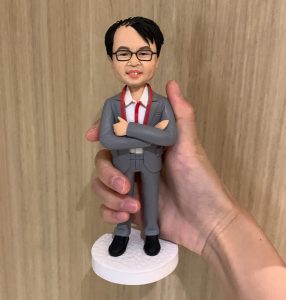 一隻單人像公仔，穿著灰色西裝、白恤衫和紅色領帶，雙手交叉抱胸，站在白色圓形底座上。背景為木紋牆面。這款人像公仔適合作為升職禮物，強調單人像公仔設計。