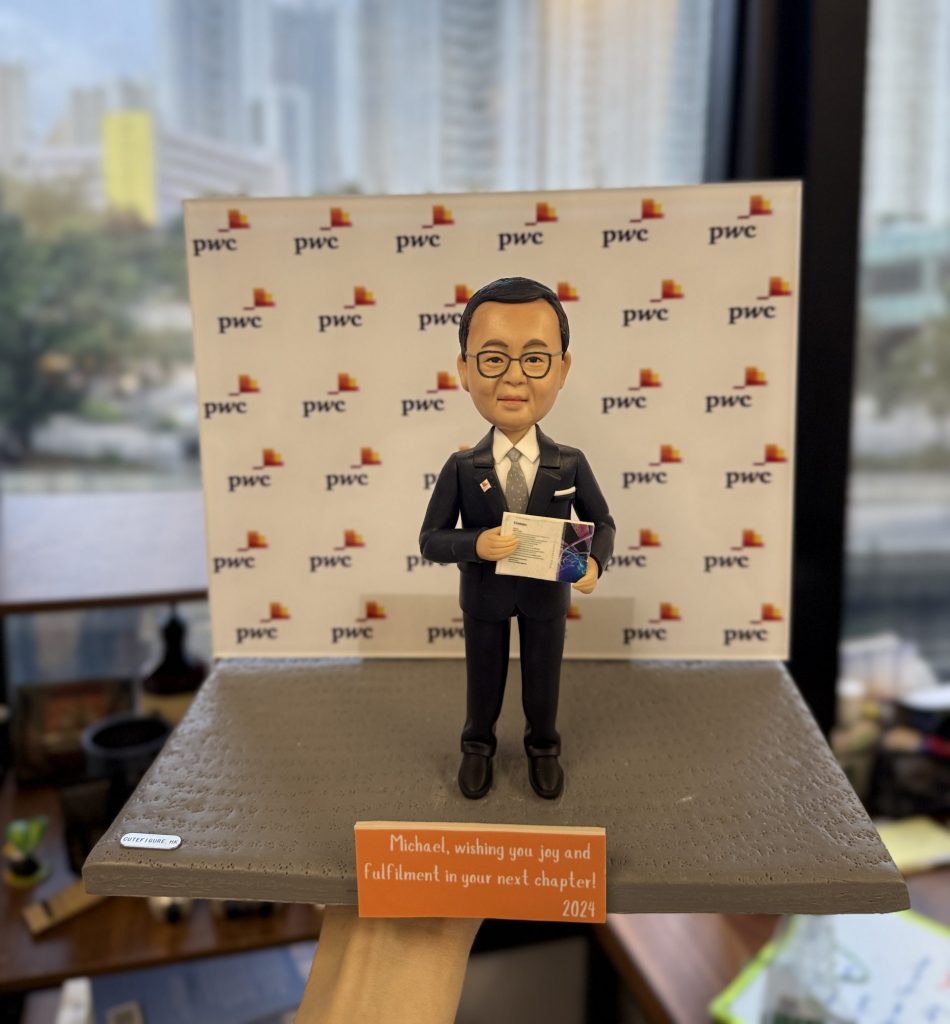 一款男性人像公仔，穿著西裝並手持文件，站立於灰色平台上，身後是印有公司LOGO（pwc）的背景牆。這款人像公仔非常適合作為升職禮物、上司禮物及公司禮物，突顯專業形象與企業歸屬感。