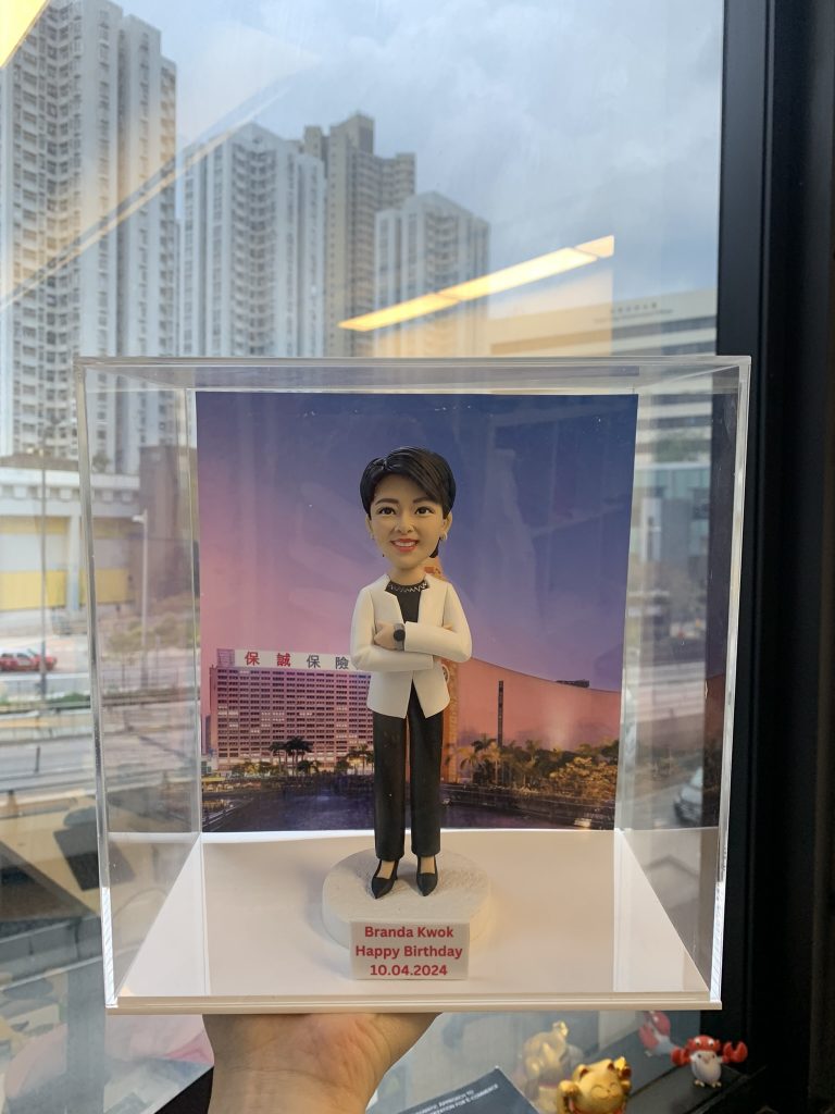 一個穿著白色西裝外套和黑色長褲的女性人像公仔，雙手交叉站立，放在透明展示盒內，背景為城市建築，底座寫有生日祝福字句。