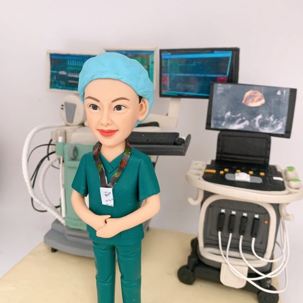 一位身穿綠色手術服、頭戴藍色手術帽的女性超聲波技師人像公仔，她自信地站在兩台高度精細的超聲波儀器模型之間，儀器上的多個顯示屏還原了掃描圖像，是一款向頂尖醫療影像專家致敬的榮休禮物。