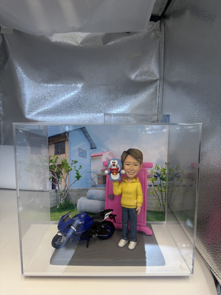 一個年輕女生人像公仔,手拿玩偶,旁邊有藍色機車和粉紅色電話亭,背景為房子和庭院。