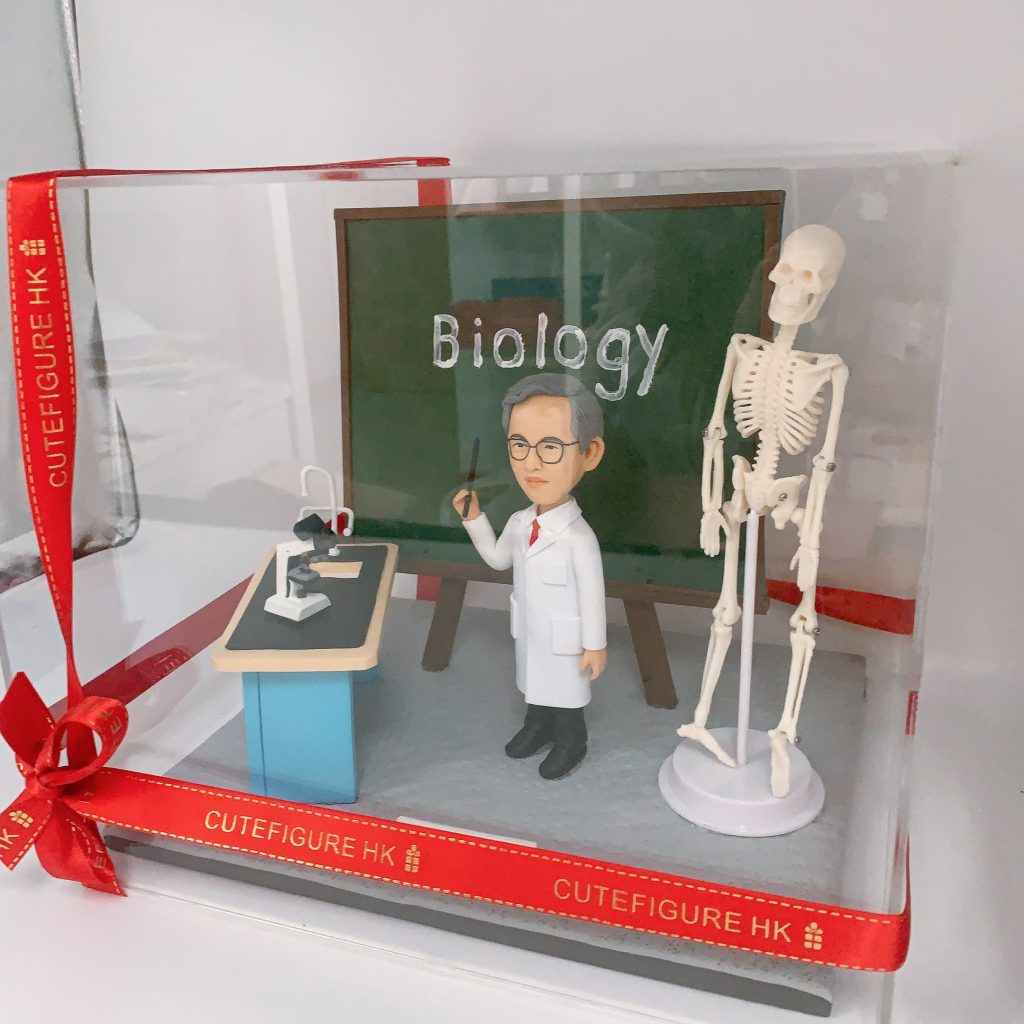 一位生物老師的人像公仔,他身穿白袍、手持教鞭,站在寫有「Biology」的黑板前,身旁還有一台顯微鏡和一具完整的人體骨骼模型,是一款極具創意的老師榮休禮物。