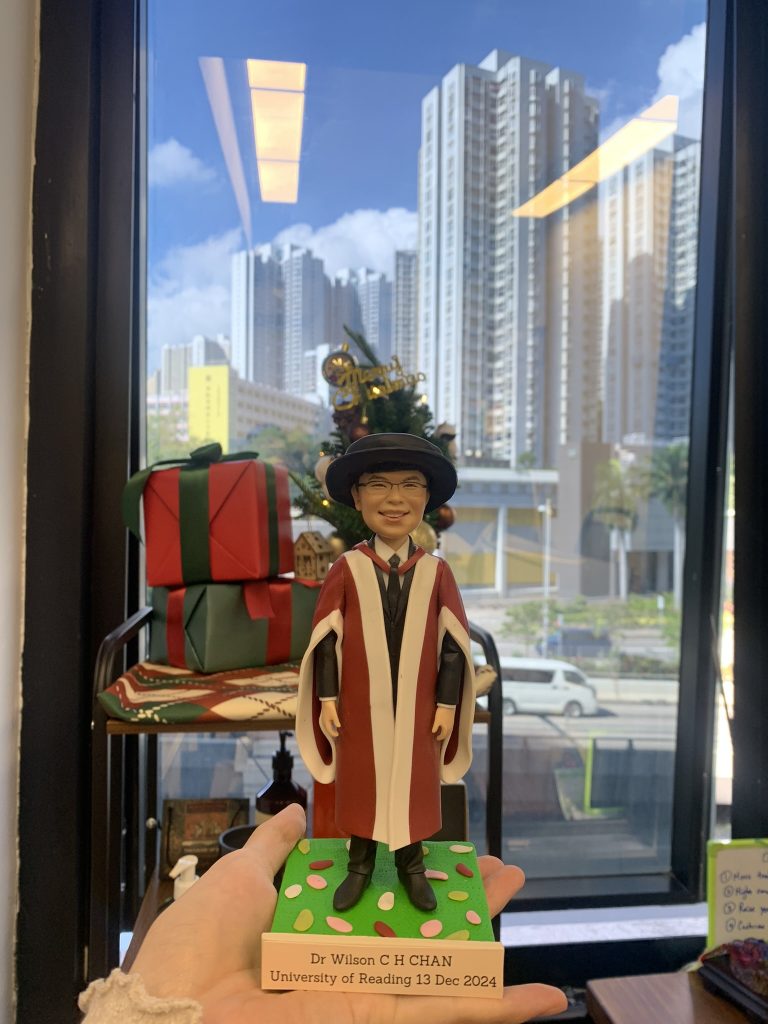 一個男人人像公仔，穿著雷丁大學的紅白博士畢業袍和黑色學位帽，戴著眼鏡，手持畢業證書，站在寫有「Dr Wilson C H CHAN University of Reading 13 Dec 2024」的底座上。
