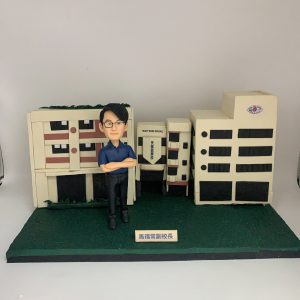 天保民學校馬福常副校長的人像公仔，他雙臂交叉，自信地站立在精細的學校建築模型前。這是一款極具份量的榮休禮物，象徵著他對學校的貢獻。