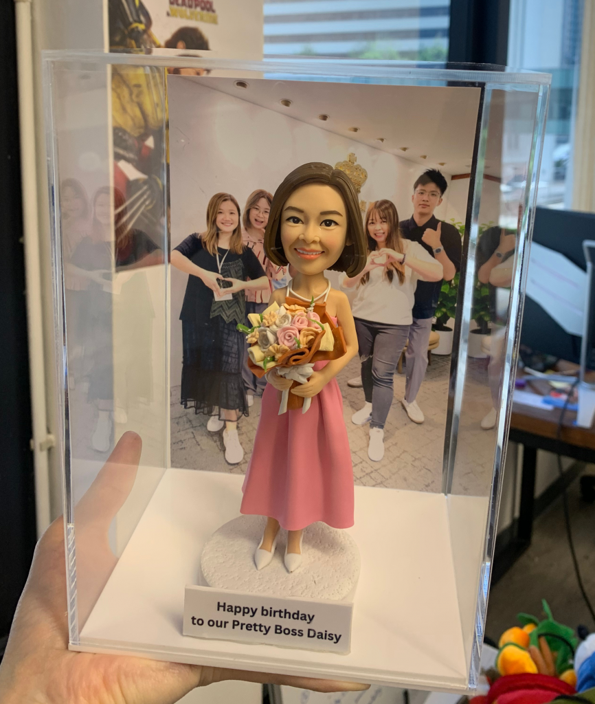 一個穿著粉紅色連身裙、雙手捧花的人像公仔，放在透明展示盒內，背景是與同事合照的相片，底座有生日祝福字牌。