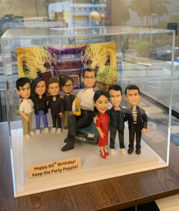 八位人像公仔站在一起合照，中央一位男士坐在車上，手持飲品，旁邊有男性及女性同事，背景是金龍裝飾的建築，底座寫有生日祝福語。