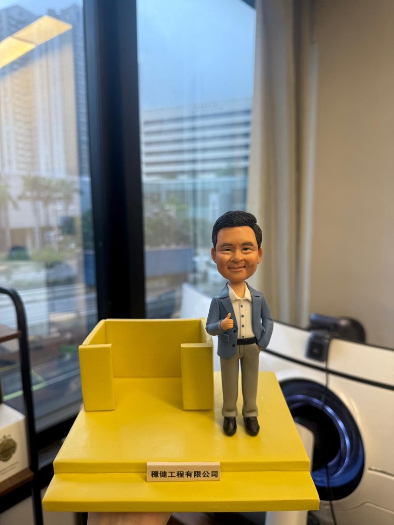 一款為「穩健工程有限公司」主管訂製的人像公仔卡片座，公仔面帶微笑、身穿藍色西裝外套並做出「讚好」手勢，站在一個鮮黃色的實用卡片座旁，是一款充滿正能量的工程師禮物或升職禮物。