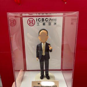 一款贈予工銀亞洲 (ICBC Asia) 員工的榮休禮物。這款人像公仔被放置在刻有「Happy Retirement 2025」的亞加力膠展示盒中，背景是工銀亞洲的公司標識。