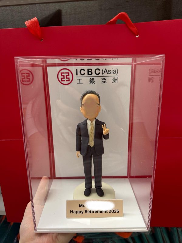 一款贈予工銀亞洲 (ICBC Asia) 員工的榮休禮物。這款人像公仔被放置在刻有「Happy Retirement 2025」的亞加力膠展示盒中，背景是工銀亞洲的公司標識。