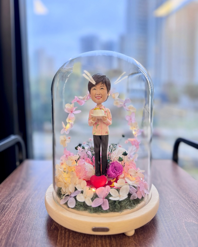 一位女性人像公仔站在花卉和燈飾環繞的玻璃罩中，手持禮物盒，背景有辦公大樓。