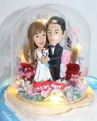 一對新人公仔穿著婚紗和西裝，置於玻璃罩內，周圍有紅色玫瑰與花卉裝飾，下方有紀念日期。